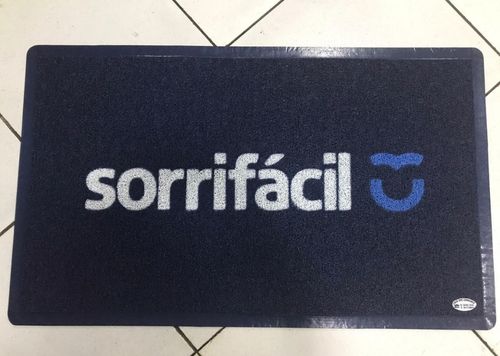 Capacho personalizado azul-marinho com logotipo Sorrifácil em branco e detalhe azul, instalado sobre piso de ladrilho branco, fotografia realista de ambiente comercial