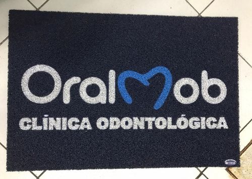 Capacho personalizado azul-marinho com logotipo OralMob Clínica Odontológica em branco e azul, instalado sobre piso de ladrilho branco, fotografia realista de ambiente comercial