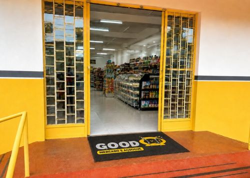 Entrada de mercado e açougue com capacho personalizado preto Good escrito em amarelo, portas de vidro amarelas, piso vermelho, prateleiras de produtos ao fundo, fotografia realista de ambiente comercial