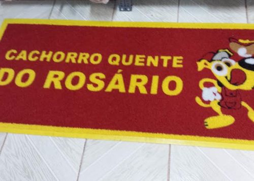 Capacho retangular vermelho e amarelo personalizado com o texto Cachorro Quente do Rosário e mascote ilustrado, sobre piso de madeira clara, fotografia realista de ambiente comercial