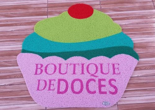 Capacho personalizado em formato de cupcake colorido com o texto Boutique de Doces em rosa, sobre piso de madeira, fotografia realista de ambiente comercial